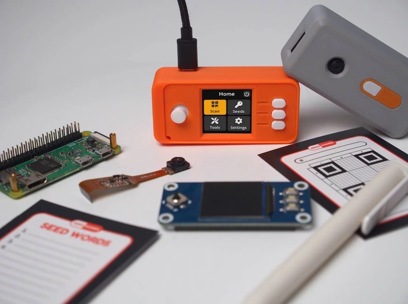 SeedSigner in an orange 3D-printed enclosure displaying the home screen, surrounded by a Raspberry Pi Zero, camera module, LCD HAT, SeedQR backup card, seed words notepad, and a gray enclosure